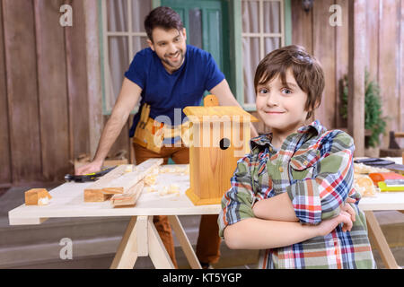 Padre Felice appoggiata al tavolo con gli attrezzi e birdhouse mentre guarda il ragazzo sorridente con bracci incrociati Foto Stock