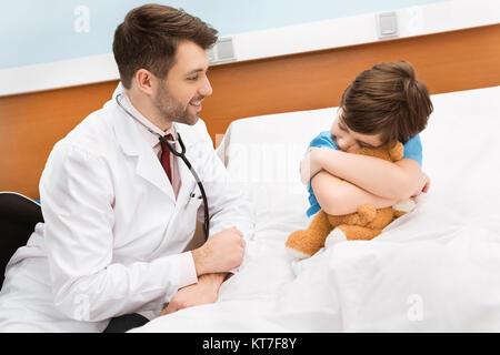 Sorridente pediatra guardando il ragazzino abbracciando Teddy bear nel letto di ospedale Foto Stock