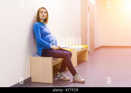 Malinconici incinta Donna seduta su una panchina all'ospedale hall e trattenere il foglio di carta. Foto Stock