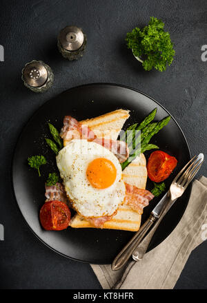 Tettuccio di tiro di prima colazione o il pranzo con uovo fritto, pane tostato, asparagi verdi, pomodori e pancetta sulla piastra nera. Foto Stock