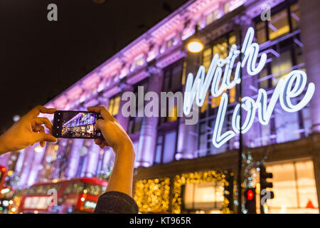 Oxford Street, Londra, 23 dic 2017. Dal grande magazzino Selfridges offre un 'con amore...dall' messaggio sulla sua facciata per Natale, per la gioia degli acquirenti di prendere un sacco di scatti delle illuminazioni. La sera prima della Vigilia di Natale, il clima mite ha portato migliaia di last minute shoppers, navigazione per occasioni e presenta, godendo le luci di Natale e passeggiando lungo i marciapiedi affollati. Credito: Imageplotter News e sport/Alamy Live News Foto Stock