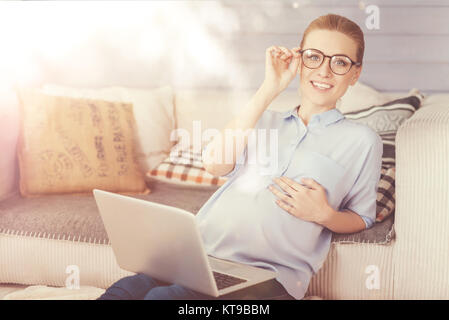 Piacevole giornata. Allegro smilign donna incinta utilizzando laptop e appoggiata sul lettino durante il riposo a casa Foto Stock