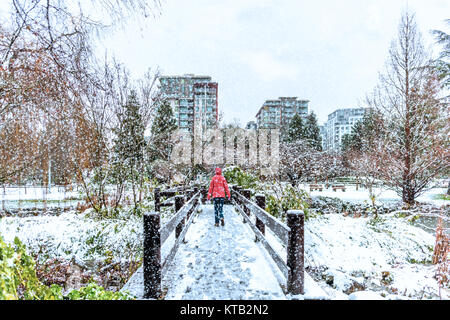 In un giorno di neve in inverno, una donna in giacca rossa passeggiate attraverso un ponte tra le coperte di neve verde di alberi e cespugli, verso moderni, molti piani di hous Foto Stock
