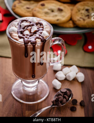 Frech cioccolata calda con una manciata di mini marshmallows e lo sciroppo di cioccolato spruzzato sulla parte superiore. Un piatto casereccio biscotti con scaglie di cioccolato è in bac Foto Stock