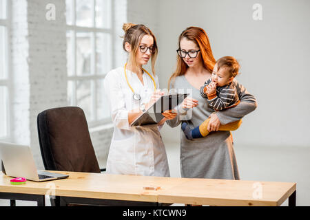 Madre con il suo bambino durante la consultazione con la giovane donna pediatra in piedi la carica bianca Foto Stock