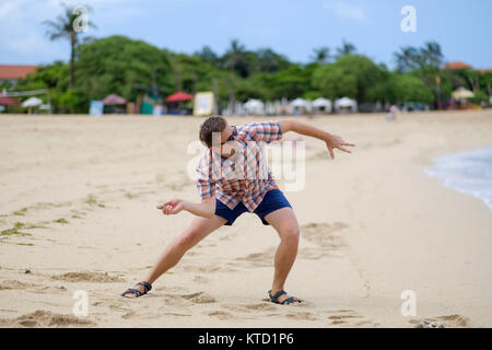 Felice uomo caucasico giocando sulla spiaggia che lanciano pietre sulla ocean, Bali Indonesia, per esterno Foto Stock