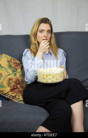 Giovane bella bionda donna caucasica con luce occhi marroni seduti sul divano a mangiare popcorn. Foto Stock