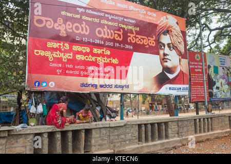 Senzatetto donna con suo Figlio seduto sul muro, grande striscione in background in Bangalore, Bengaluru, Karnataka, India, Asia. Foto Stock