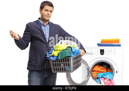 Infelice ragazzo elegante tenendo un cesto per la biancheria riempito con vestiti nella parte anteriore di una macchina di lavaggio isolata su sfondo bianco Foto Stock