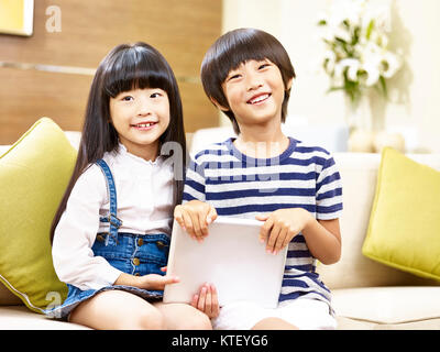 Due graziosi bambini asiatici little boy e poco ragazza seduta sul lettino azienda digitale compressa guardando sorridente della fotocamera Foto Stock