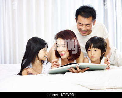 Felice famiglia asiatica con due bambini che giace sulla parte anteriore a letto a leggere un libro insieme. Foto Stock