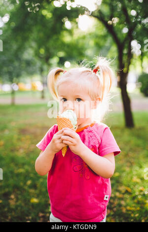 Poco funny girl bionda mangiare blu dolci gelato in una cialda cup su un verde sfondo estiva nel parco. cosparso il suo viso e le guance e risate. Foto Stock