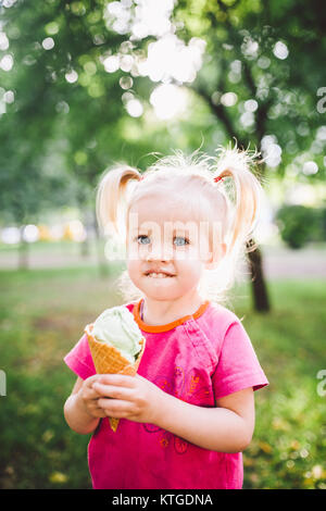 Poco funny girl bionda mangiare blu dolci gelato in una cialda cup su un verde sfondo estiva nel parco. cosparso il suo viso e le guance e risate. Foto Stock