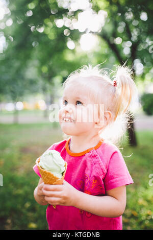 Poco funny girl bionda mangiare blu dolci gelato in una cialda cup su un verde sfondo estiva nel parco. cosparso il suo viso e le guance e risate. Foto Stock