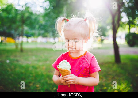 Poco funny girl bionda mangiare blu dolci gelato in una cialda cup su un verde sfondo estiva nel parco. cosparso il suo viso e le guance e risate. Foto Stock