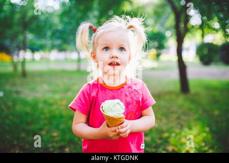 Poco funny girl bionda mangiare blu dolci gelato in una cialda cup su un verde sfondo estiva nel parco. cosparso il suo viso e le guance e risate. Foto Stock