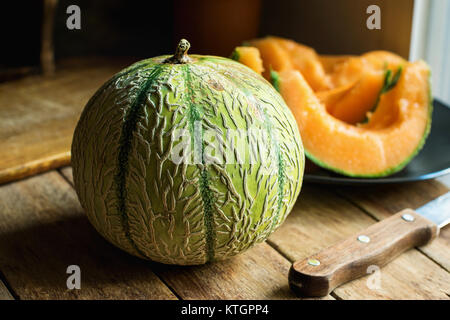 Intero e mature a fette di melone organico con succosa polpa arancione sulla piastra. Il taglio della lama di bordo su tavola di legno. Cucina rustica interno accogliente atmosfera Foto Stock