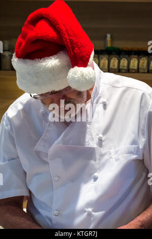 Uomo anziano con la barba in chef bianchi e un cappello da Babbo Natale preparare il cibo in una cucina a tempo di Natale Foto Stock