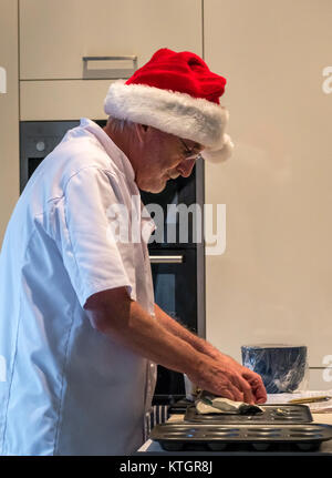 Uomo anziano con la barba in chef bianchi e Santa hat preparazione e ingrassaggio tritare teglie di torta in una cucina per la preparazione delle torte di carne macinata a Natale Foto Stock