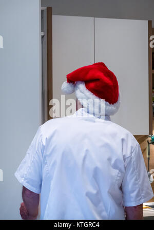 Uomo anziano in chef bianchi e Santa hat guardando in un frigorifero in una cucina a tempo di Natale Foto Stock