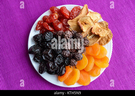 Un mucchio di frutta secca (mele, prugne, albicocche, pere, mirtilli) su una piastra bianca su uno sfondo viola. Mix di antiossidante. Un prodotto utile. Prop Foto Stock