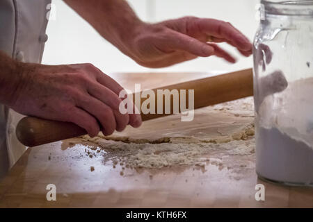 Close up dei più vecchi in chef bianchi pasta di laminazione con un matterello facendo sfoglia su un blocco di legno con un vasetto di farina in una cucina Foto Stock