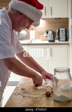 Uomo anziano in chef bianchi e Santa hat pasta di rotolamento con il mattarello rendendo la pasta per torte di tritare sul blocco di legno in una cucina a tempo di Natale Foto Stock