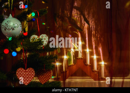 Incandescente menorah sul davanzale situato dietro le tende con l'albero di Natale in primo piano, decorate con giocattoli. La messa a fuoco su la menora Foto Stock