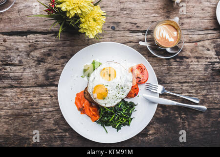 La prima colazione con uova fritte, salmone, avocado, pomodoro cotto al forno, toast e verdi. Tazza di cappuccino di aroma di legno sfondo testurizzato, laici piatta Foto Stock