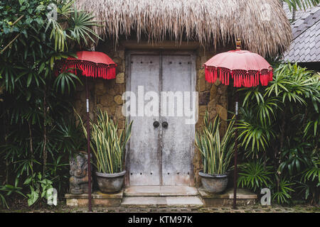 Ingresso in bali tradizionale casa con porta in legno e tetto di paglia e ombrelloni rosso Foto Stock