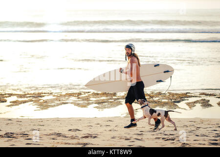 Surfer in reef stivali con tavola da surf e casco in esecuzione jogging sulla spiaggia con il suo cane al tramonto a Bali, Indonesia. Foto Stock