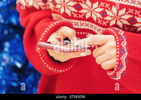 La pubblicità di natale il concetto di shopping. donna tenendo il telefono cellulare e toccando lo schermo vuoto ad albero di natale luci. indossa un maglione rosso con snowfl Foto Stock