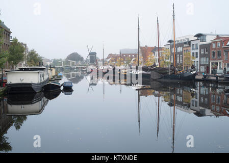 Navi, mulino a vento e case sono riflesse in un canale su una mattinata nebbiosa di Leiden, Paesi Bassi Foto Stock