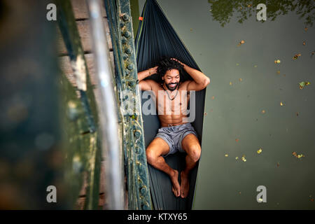 Un uomo si rilassa in una amaca espulso da un ponte su un fiume. Foto Stock