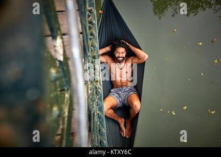 Un uomo si rilassa in una amaca espulso da un ponte su un fiume. Foto Stock