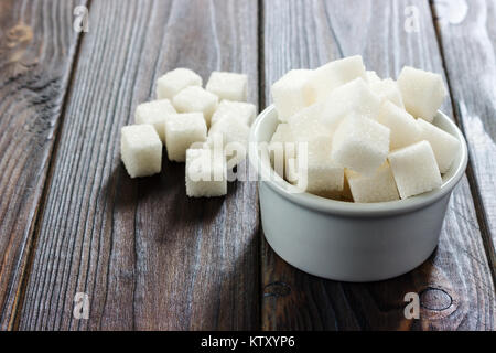 Zucchero bianco in vaso su sfondo di legno. Messa a fuoco selettiva, orizzontale. Alcuni cubetti di zucchero sono vicino al bicchiere pieno con zucchero bianco. Aspirazione di bad calor Foto Stock