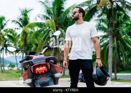 Uomo in piedi vicino alla sua moto con casco su Palm Background Foto Stock