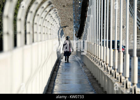 Una donna che porta una grande borsa attraversando a piedi il ponte sospeso di Clifton a Bristol, Regno Unito. Una fredda giornata invernale e vi è il ghiaccio e la neve sul sentiero. Foto Stock