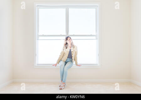 Una giovane donna seduta sul davanzale in bianco e luminoso vuoto, camera pulita sul tappeto di nuova e moderna casa, casa, appartamento in cerca di sole finestre di grandi dimensioni Foto Stock