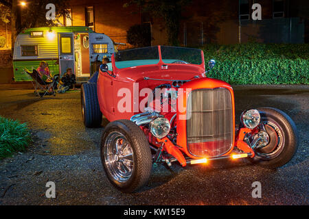 1932 red Ford hot rod roadster di notte con rimorchio camper e persone rilassante in background durante la notte. Foto Stock