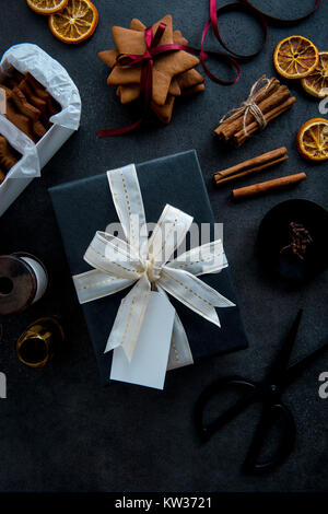 La confezione regalo per Natale. Vista superiore della tabella con il regalo in elegante carta nera con bianco e oro nastro con etichetta. Materiali e accessori. Foto Stock
