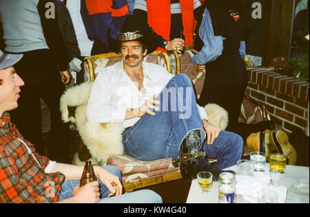 Uomo in cappello da cowboy e speroni, con una camicia bianca parzialmente rimosse, si siede con i piedi su un tavolo e di parlare e di bere alcool con un uomo che indossa una camicia scozzese, 1975. Foto Stock