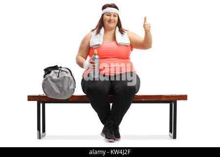 Il sovrappeso donna seduta su una panchina di legno accanto a una borsa sportiva rendendo un pollice in alto segno isolato su sfondo bianco Foto Stock