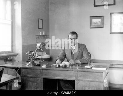 Un uomo con capelli corti, round occhiali una tuta conservatore e a poppa con un espressione del viso si siede dietro una grande scrivania di legno nel suo ufficio, con un piccolo lavabo, un inchiostro blotter, due campane, una scrivania, una lampada e un wind-up telefono visibile, Ungheria, 1929. Digitalizzate da un processo di gelatina lastra di vetro negativo. Foto Stock