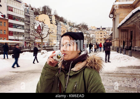 Giovane donna di bere dal bicchiere con acqua minerale terapeutica ad una sorgente calda naturale a Karlovy Vary durante il periodo invernale, Repubblica Ceca Foto Stock