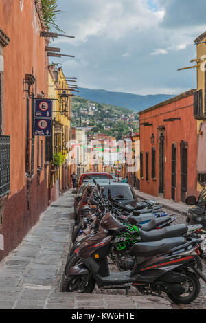 Una strada di pietra in San Miguel De Allende, con i suoi edifici colorati, foderata con i motocicli e automobili, montagne sullo sfondo e un cielo drammatico Foto Stock