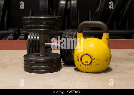 Due vecchi e utilizzato la palestra in metallo nero manubri con grande kettlebell giallo su un pavimento di legno Foto Stock