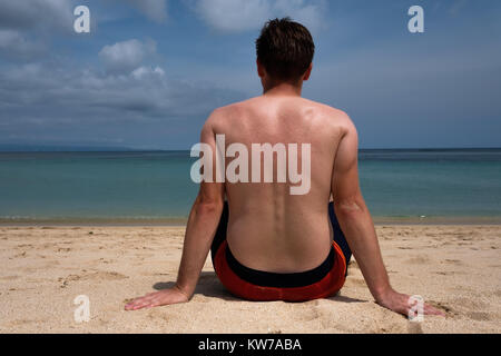 Un giovane uomo con un divertente tan da una T-shirt è in piedi sull'oceano e guarda al mare azzurro e limpido. Foto Stock