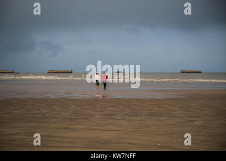 Camminando lungo la spiaggia di Arromanches les Bains con resti di cassoni di Phoenix in background. Foto Stock
