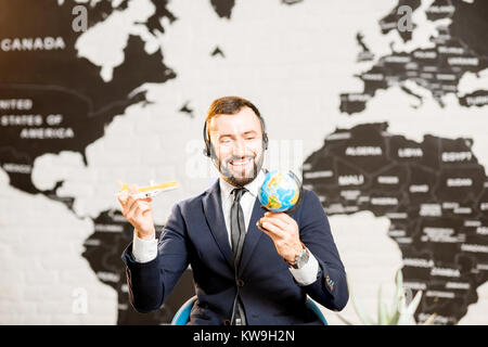 Bello agente maschio giocando con il globo e aeroplano giocattolo presso l'agenzia viaggi ufficio con mappa del mondo sullo sfondo Foto Stock